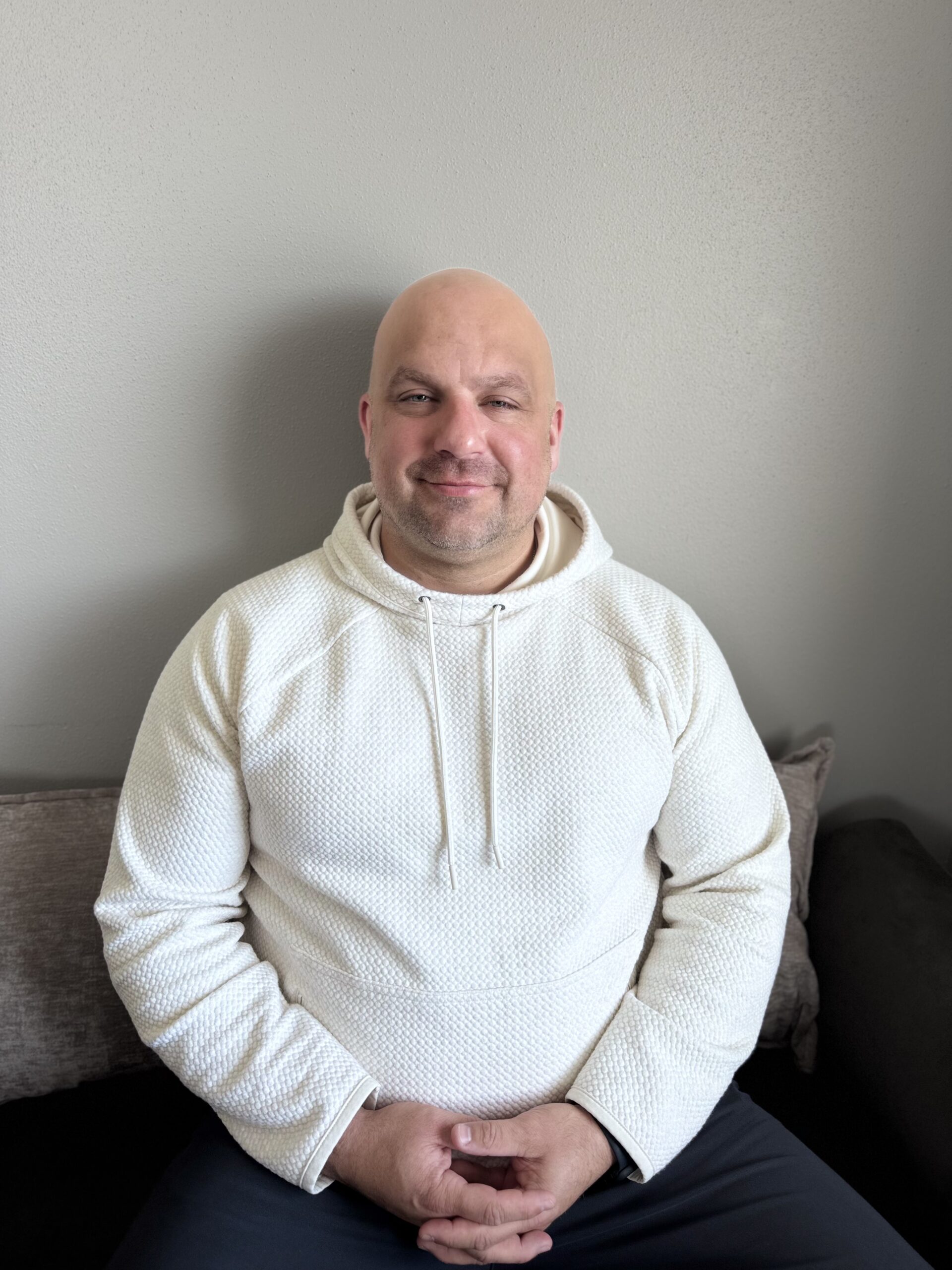 A smiling man in a white hoodie sitting on a couch against a plain wall.