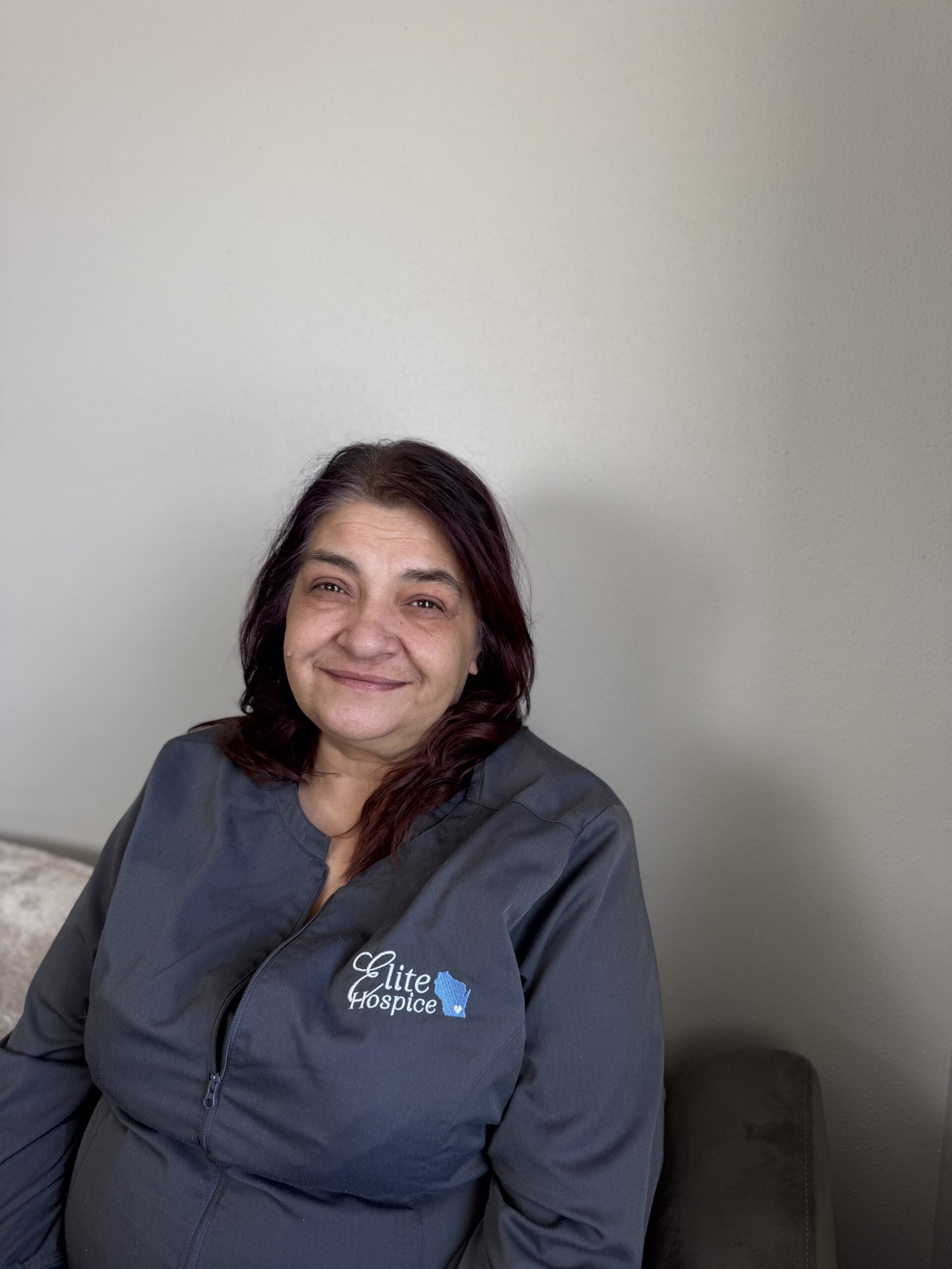 A woman smiling warmly in a gray shirt against a plain wall.